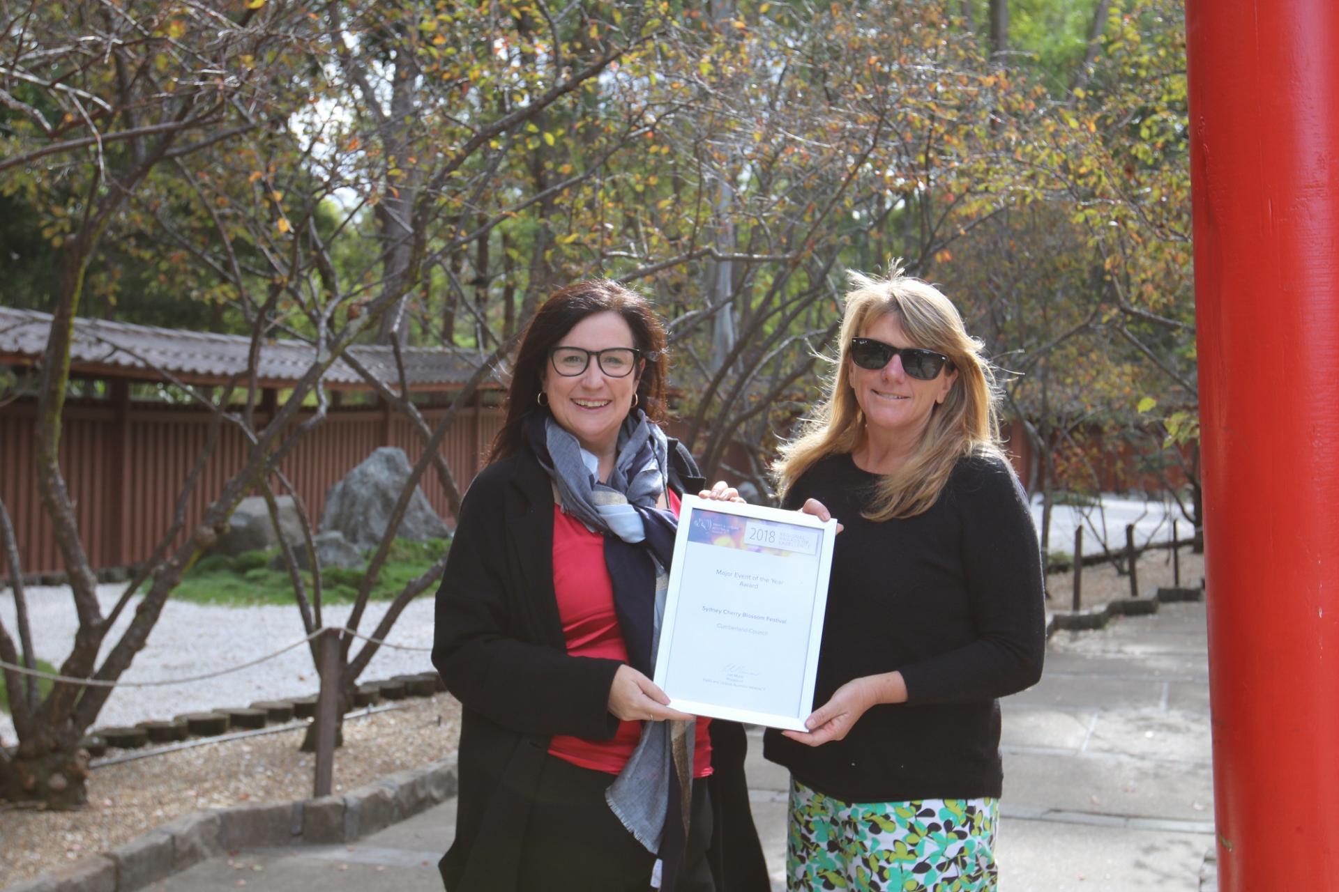 two women holding a certificate together with gardens in the background