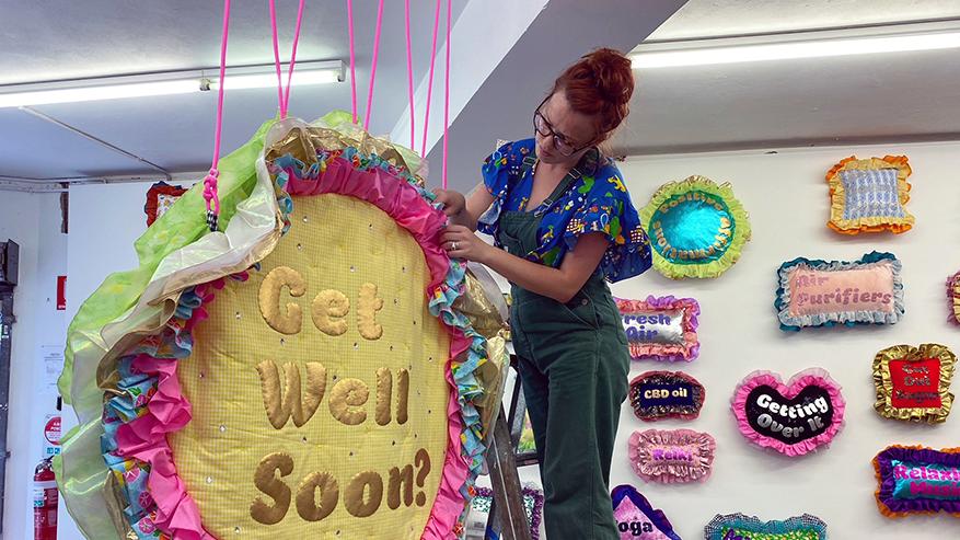 A Caucasian women with red hair is wearing bottle green overalls and brightly patterned shirt. She is standing on a ladder in a gallery installing textiles based work, including a large round colourful material rug with text on it reading ‘ Get Well Soon’. Behind her is a wall full of colourful pillows with differing text on them, including Getting over it, Mindfullness and CBD oil amongst others.