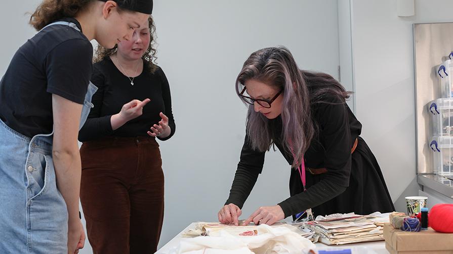 Three Caucasian women stand around a studio workshop table looking at colourful textiles based materials. One of the women is showing how to sew with red thread on calico material.
