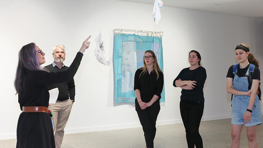 A group of people including four women and a man stand around a delicate small white hanging textiles artwork in a gallery. One of the women dressed in black with a brown belt and purple hair is pointing to the artwork, signing in Auslan to the group of people.