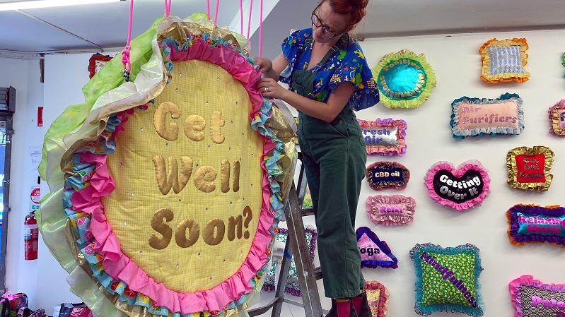 Image of Amy Claire Mills, female standing on ladder, hanging colourful textile artwork in a gallery