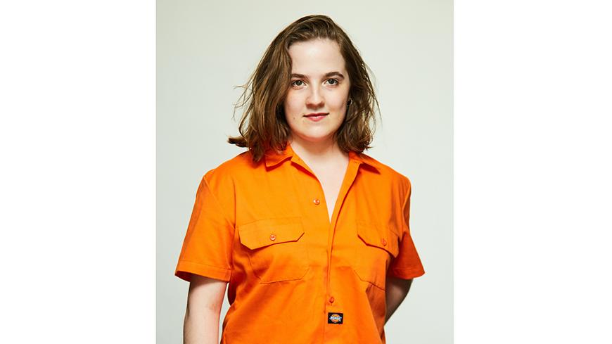 Image of Bedelia Lowrencev, female standing with shoulder length light brown hair, orange shirt and jeans
