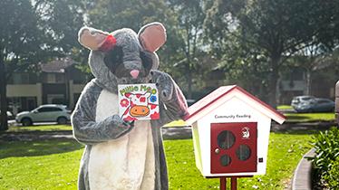 Poppy the Possum holds a book next to a Community Reading Box
